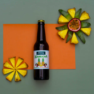 A glass bottle of Daissy pineapple passionfruit kombucha surrounded by slices of fresh pineapple and cut passionfruit